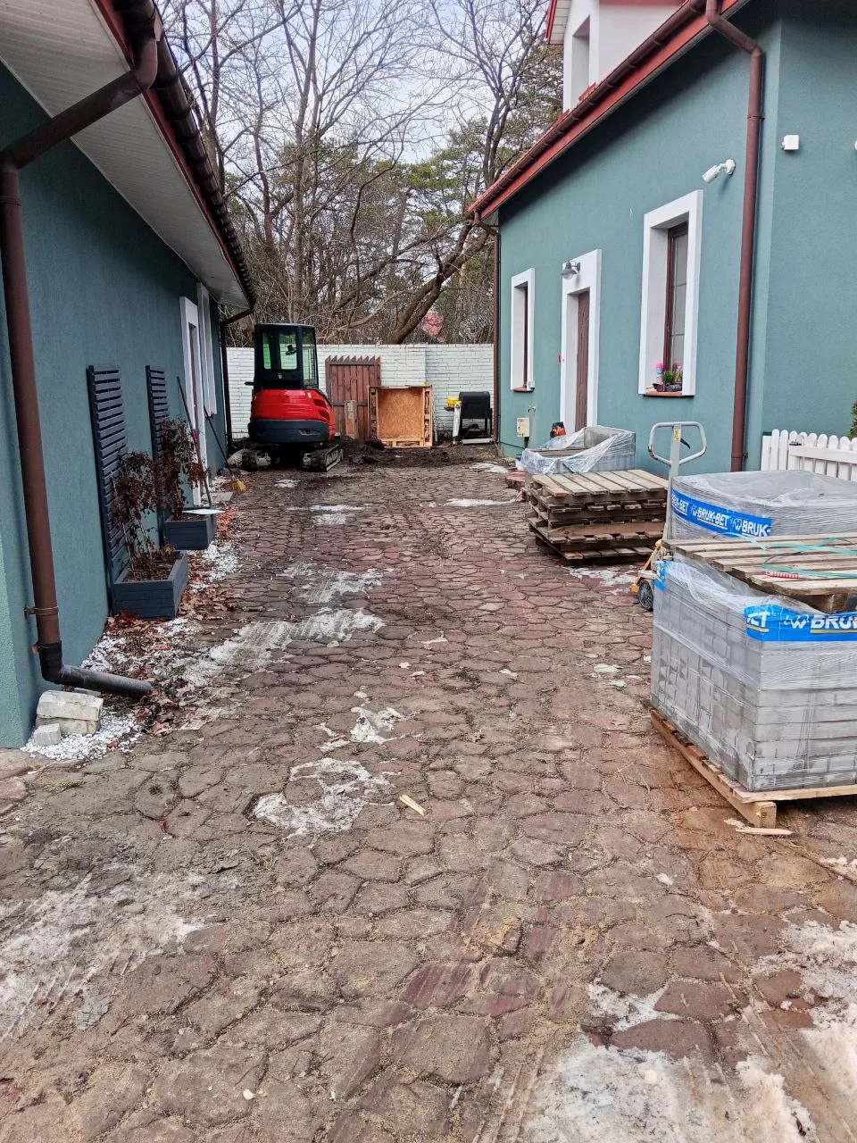 Układanie kostki brukowej przy budynku, widoczne maszyny oraz materiały budowlane